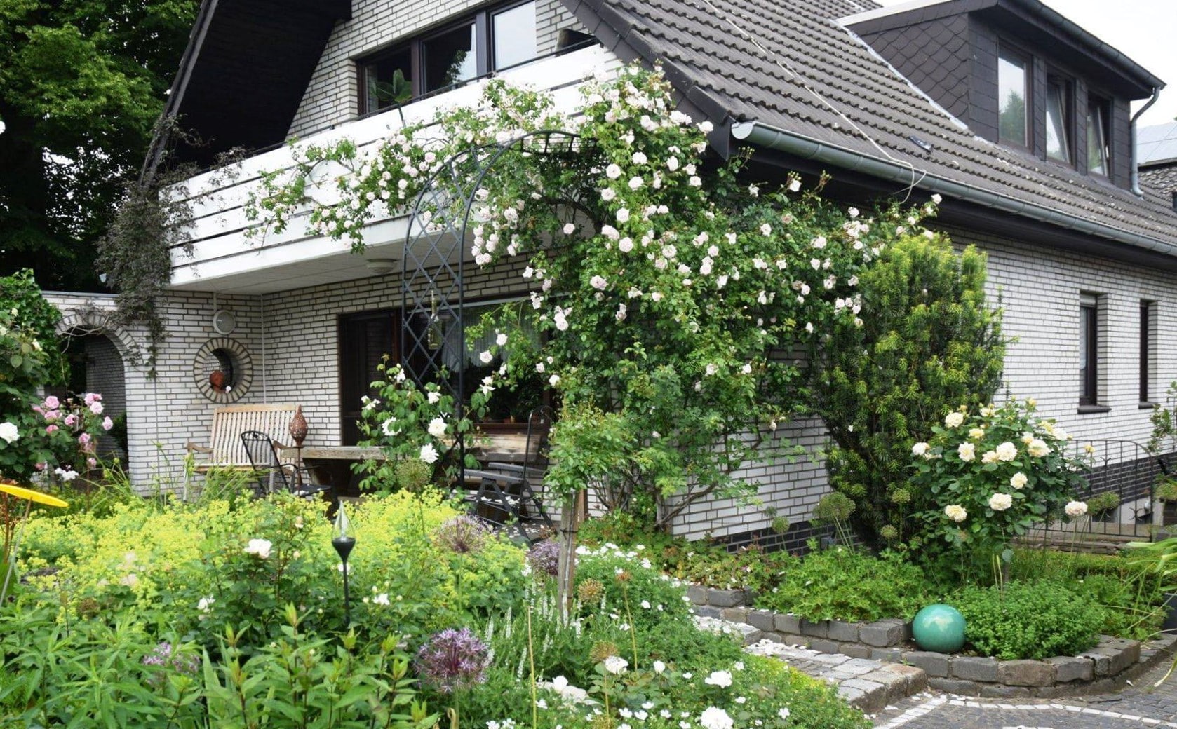 Halbbogen Bagatelle mit weißer Kletterrose markiert Übergang von Terrasse am Haus zum Garten