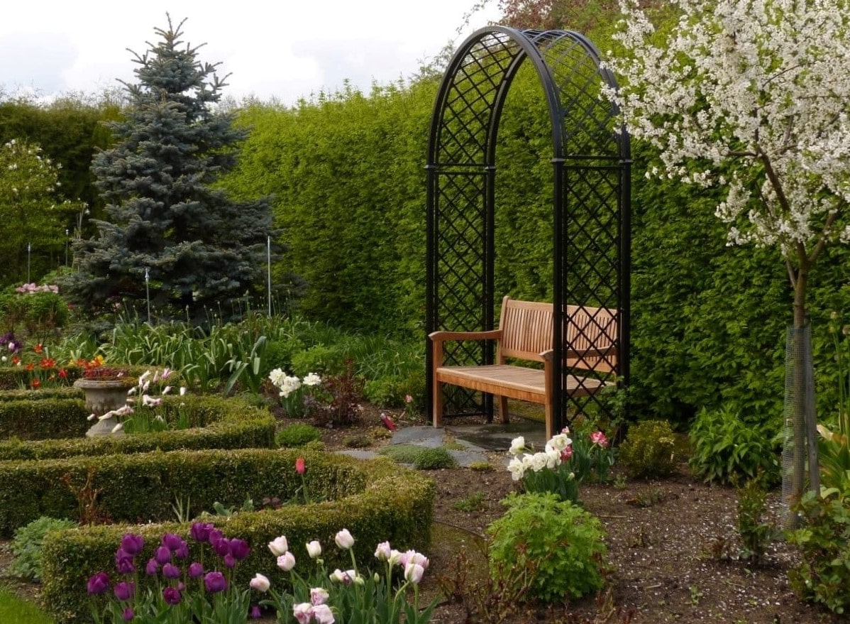 Frühlingsgarten mit Metall Rosenbogen Portofino von Classic Garden Elements und Holzbank vor hoher Hecke