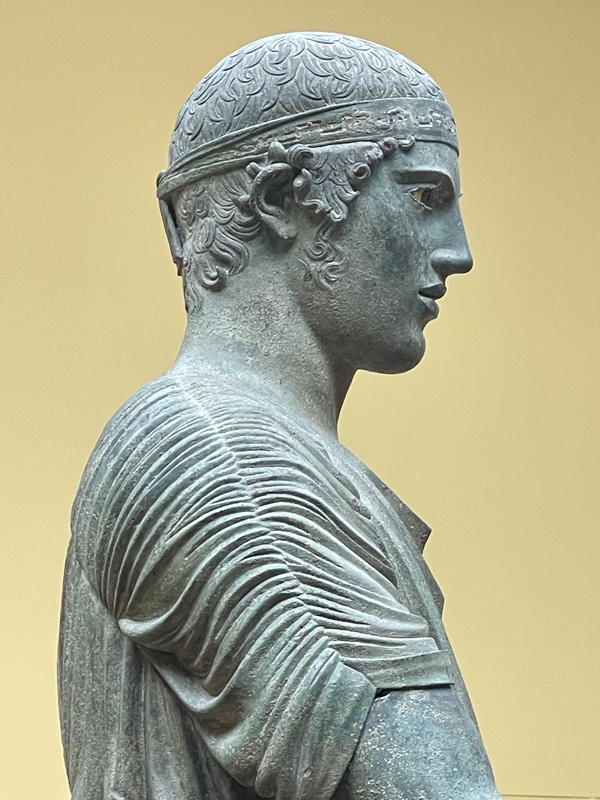 Wagenlenker von Delphi Detail des Kopfes mit Haaren und Stirnband sowie Schultern mit Tunika Wagenlenker von Delphi Detail des Kopfes mit Haaren und Stirnband sowie Schultern mit Tunika